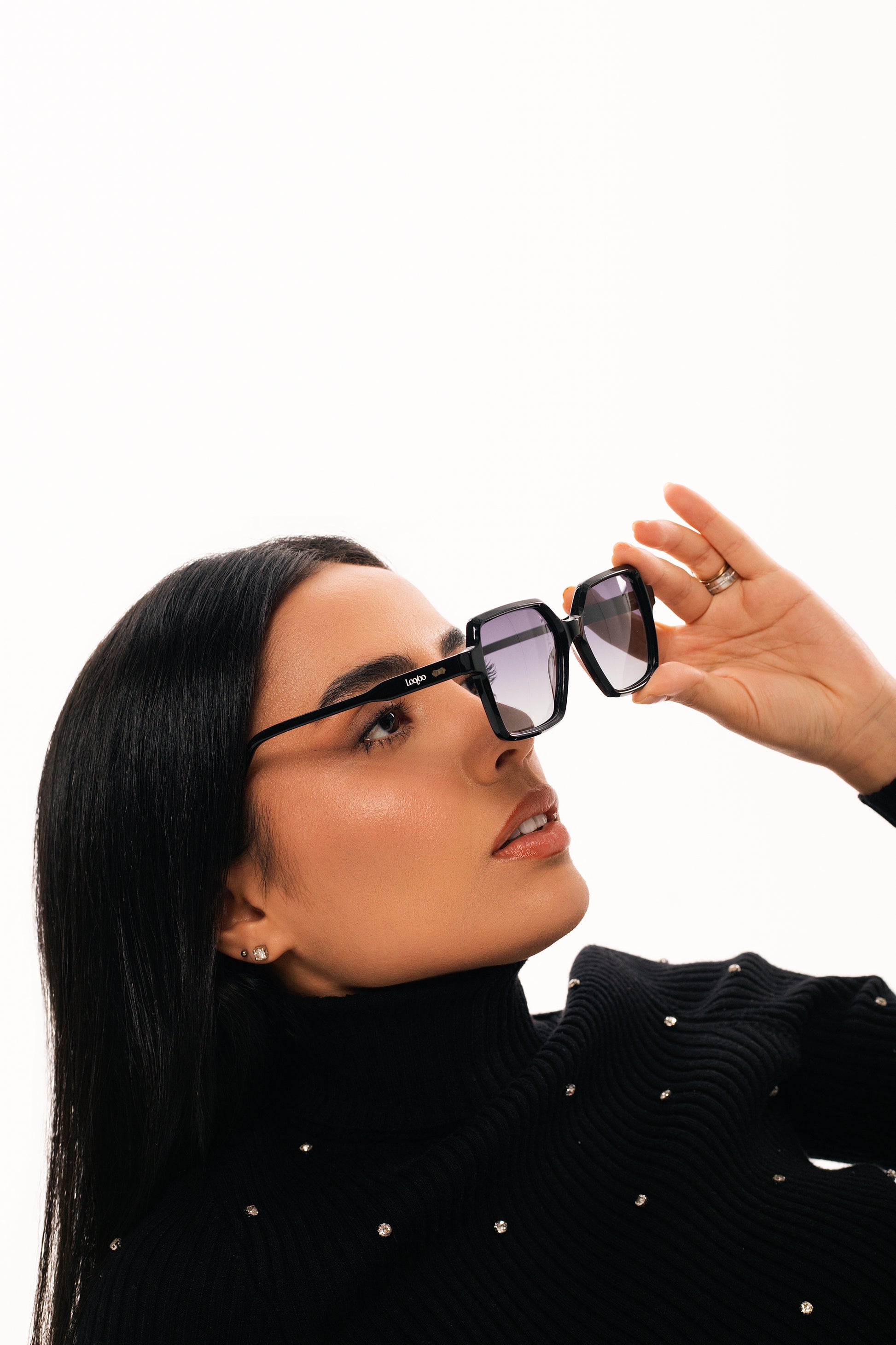 Woman wearing black sweater, holding a pair of black acetate frame sunglasses with gradient lenses, on a white background.