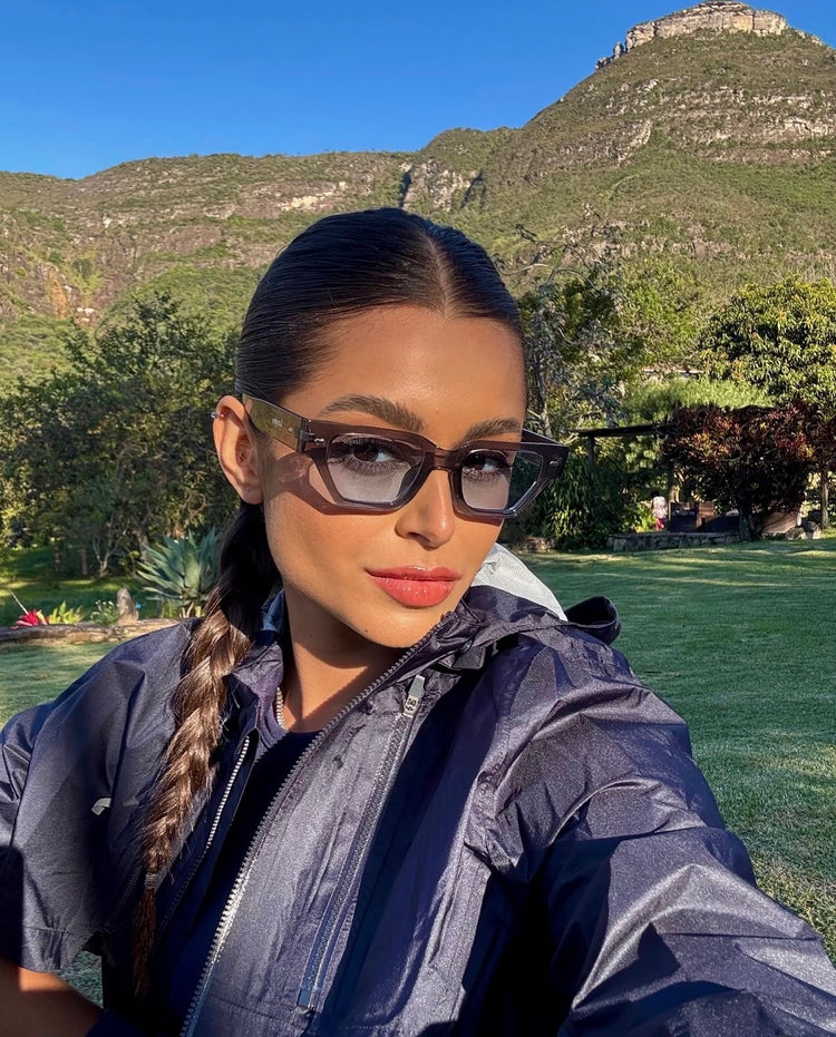 Woman wearing angular sunglasses outdoors with mountains and greenery in the background.