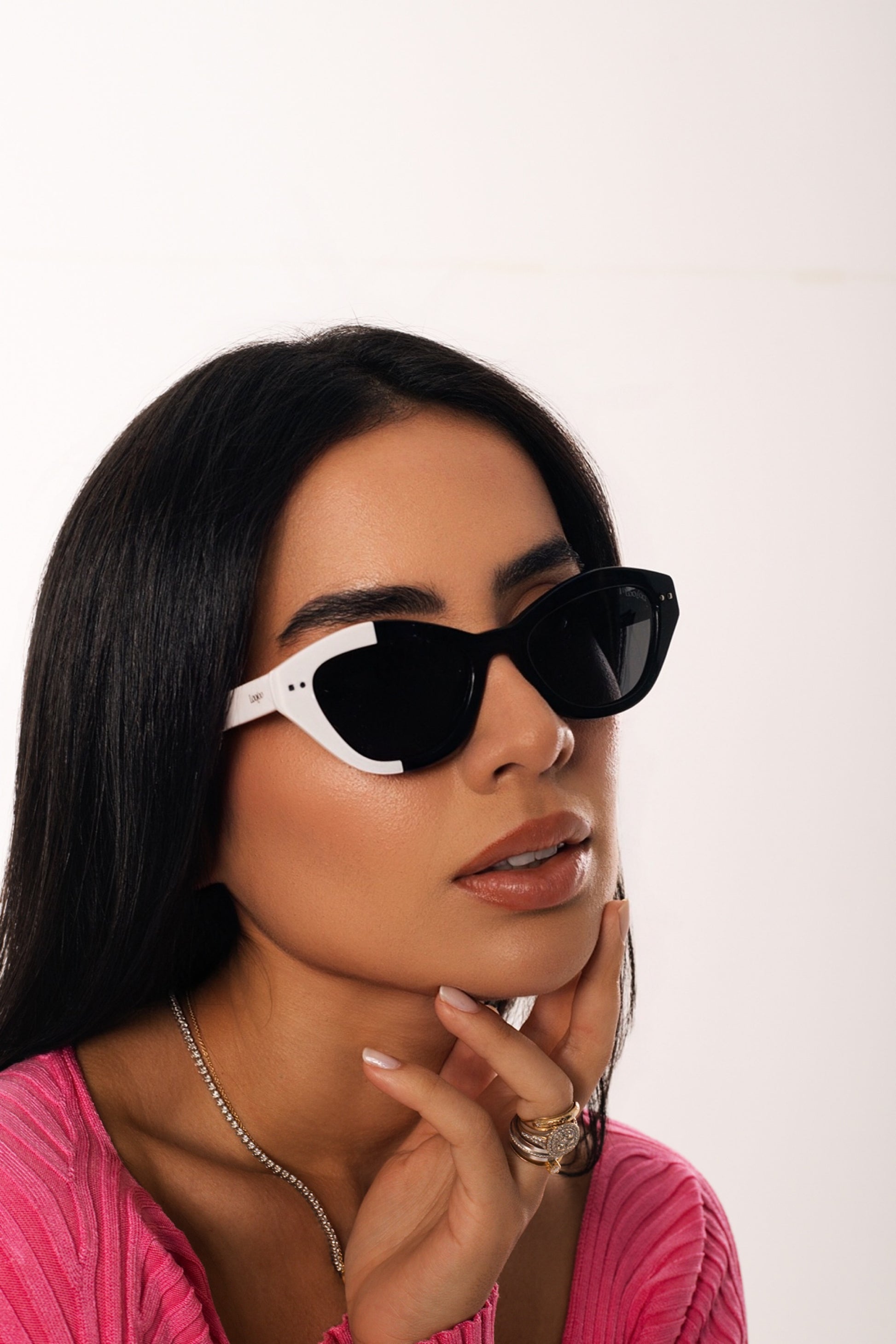 Woman wearing pink shirt, wearing a pair of black and white acetate frame sunglasses, on a white background.