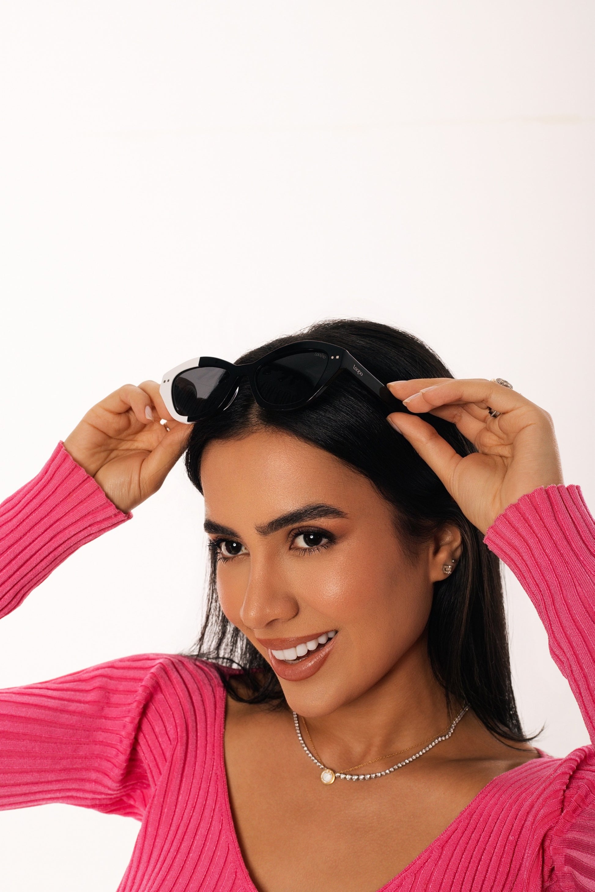 Woman wearing pink shirt, wearing a pair of black and white acetate frame sunglasses, on a white background.