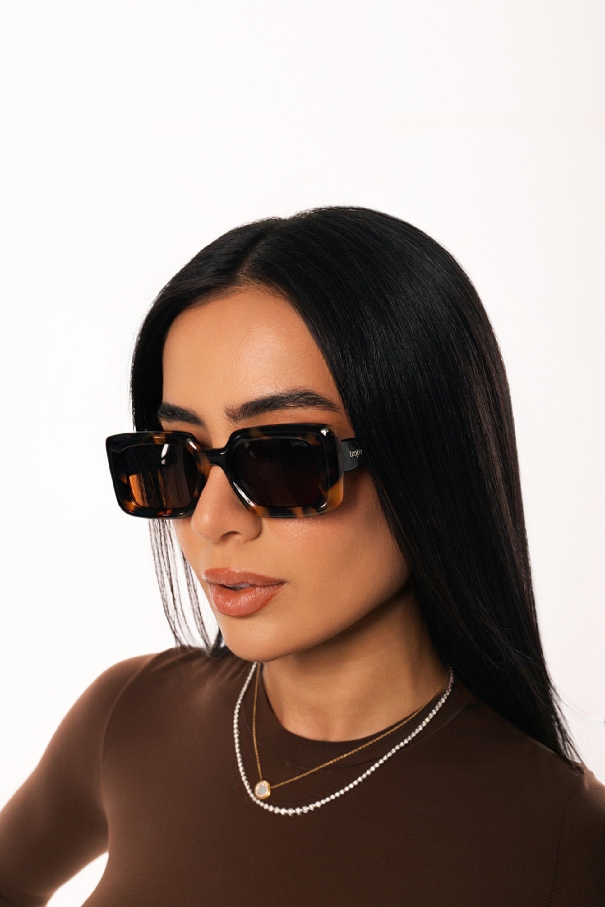 Woman wearing brown outfit, wearing a pair of tortoise acetate frame with brown lenses, on a white background.