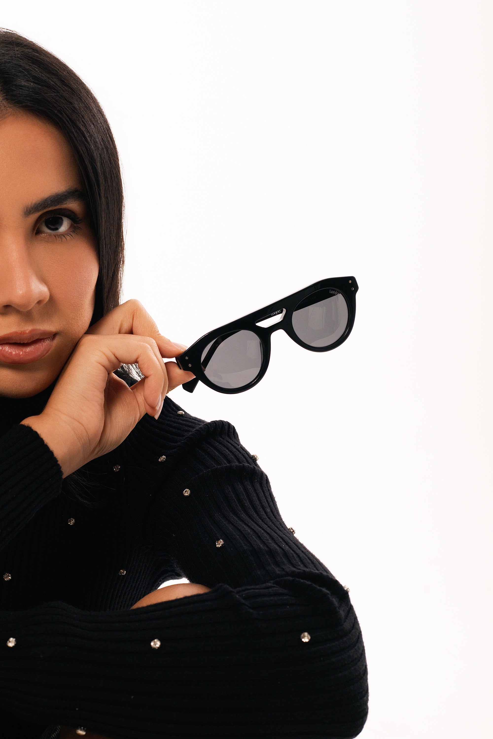 Woman wearing black sweater, holding a pair of black acetate frame  sunglasses, on a white background.
