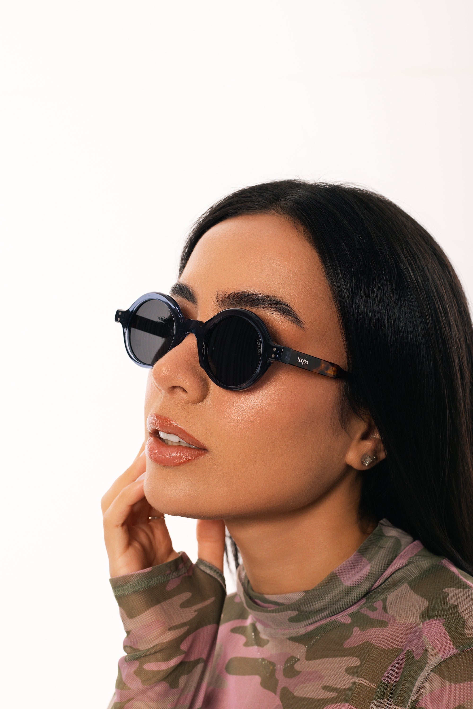 Woman wearing camouflage shirt, wearing a pair of blue and tortoise acetate frame sunglasses, on a white background.