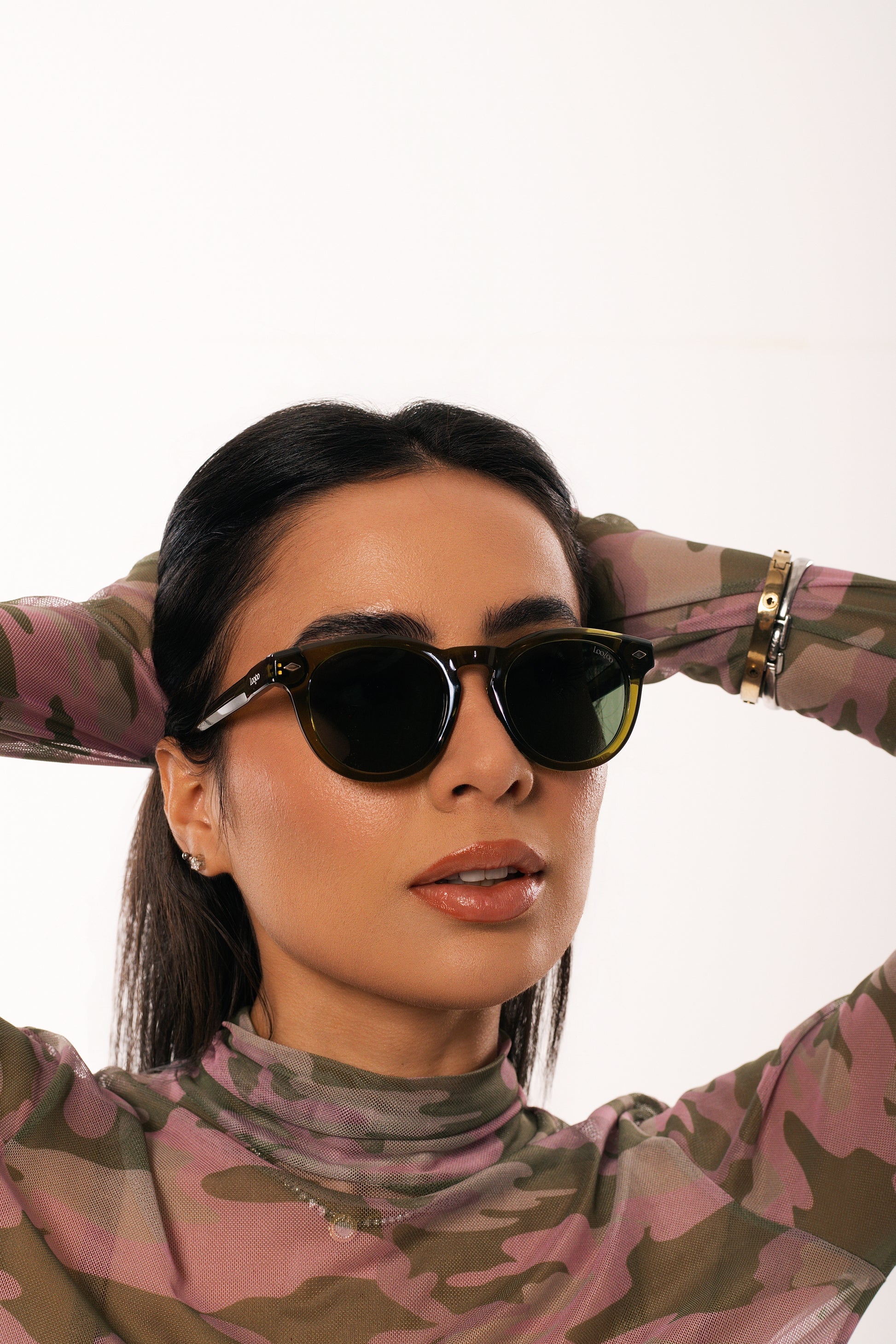 Woman wearing camouflage shirt, holding a pair of green acetate frame and green lenses sunglasses, on a white background.