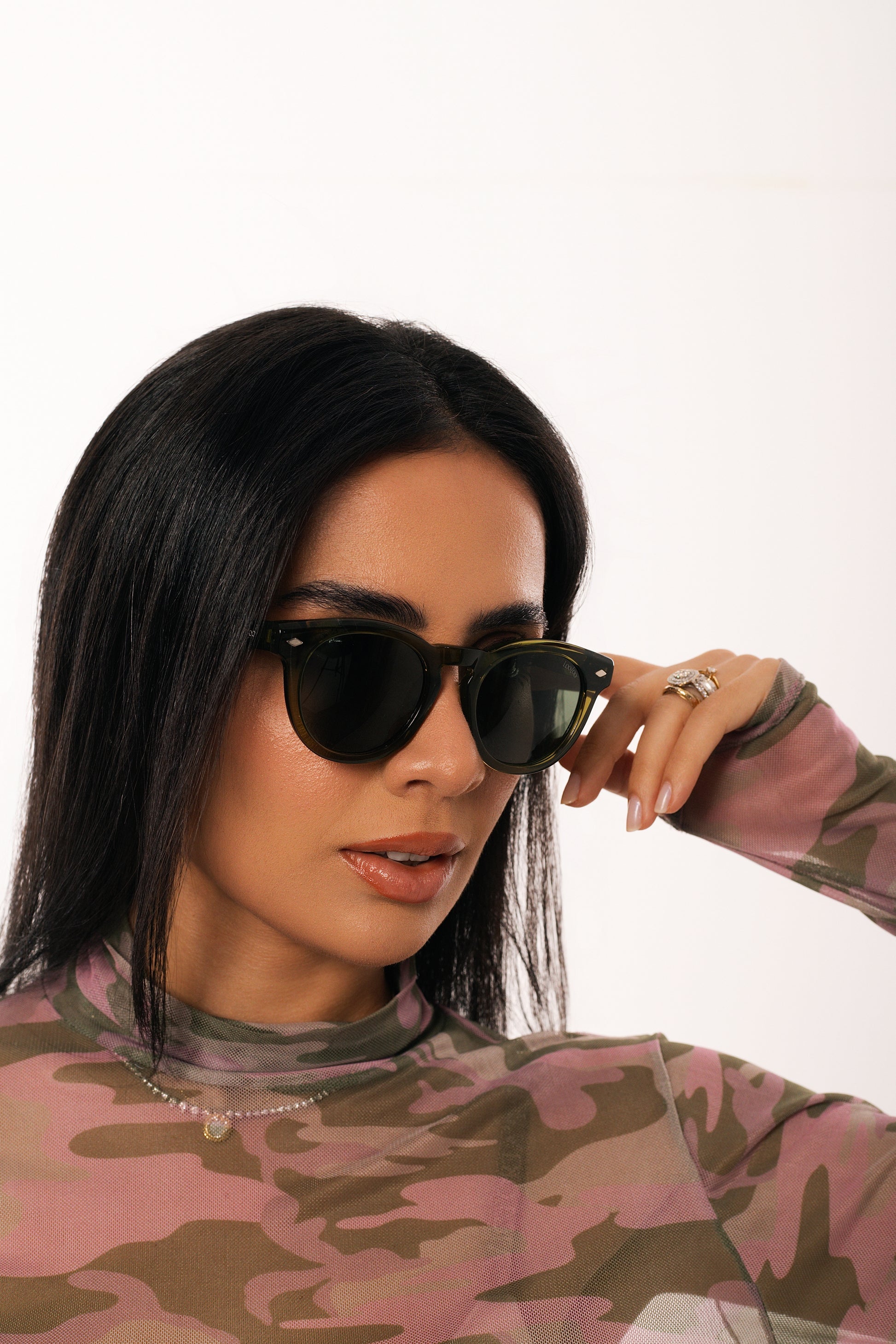 Woman wearing camouflage shirt, wearing a pair of black acetate frame sunglasses, on a white background.